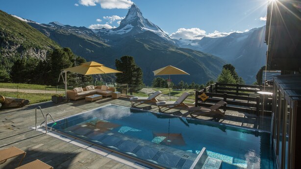 Ein Poolbereich mit Liegestühlen und Sonnenschirmen, Blick auf Berge und ein klarer Himmel.