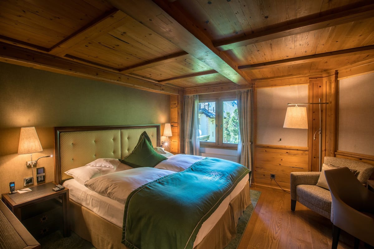 A hotel room with a wooden ceiling, a bed with green bedding, a bedside table with a lamp, and a chair.