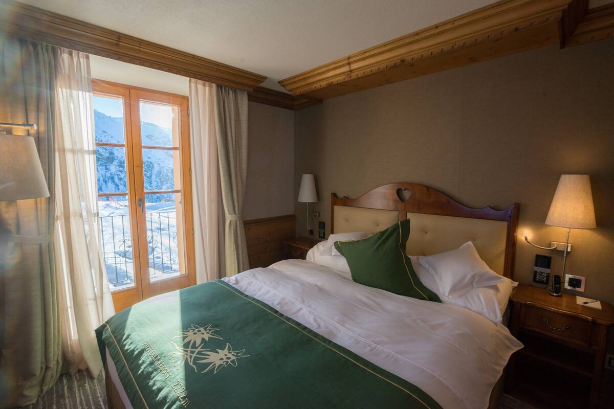 A hotel room with a bed, a nightstand, a lamp, and a view of mountains through a window.