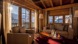 Ein gemütliches Zimmer mit Holzbalkendecke, großen Fenstern mit Blick auf die Berge, einer Couch, einem Couchtisch und einer Lampe.