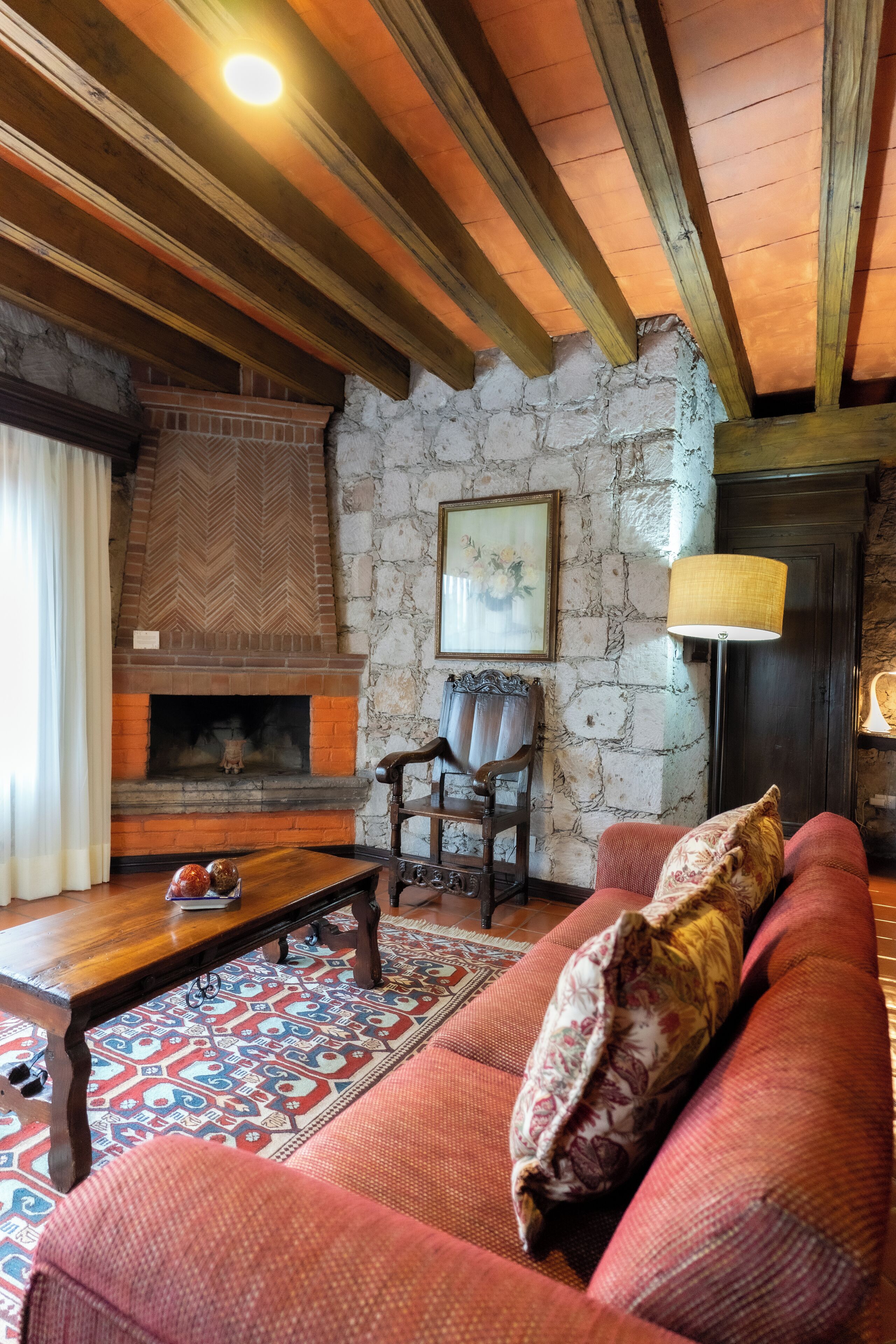 A cozy living room with a fireplace, wooden beams, a patterned rug, and a red sofa.