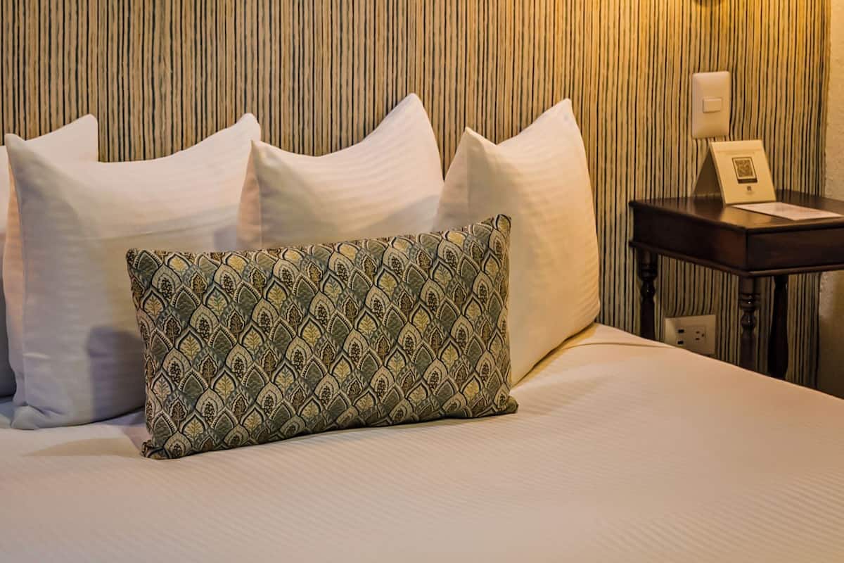 Una cama bien hecha con almohadas blancas y estampadas, una lámpara fijada a la pared y una mesita con un marco de fotos y un libro.