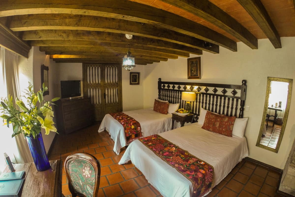 Una habitación con dos camas, techo de madera, una mesita con una lámpara y una silla.