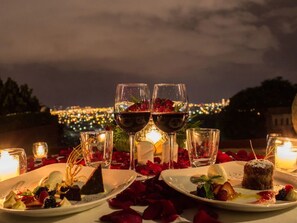 Una cena romántica con copas de vino, pétalos de rosa y platos de postre sobre una mesa con vistas a la ciudad.