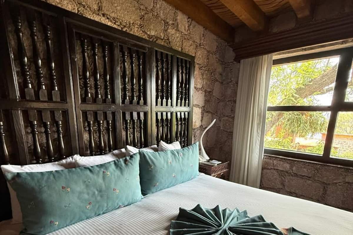 Una habitación con pared de piedra, techo de madera, cama con colcha a rayas y ventana con vista a la vegetación.