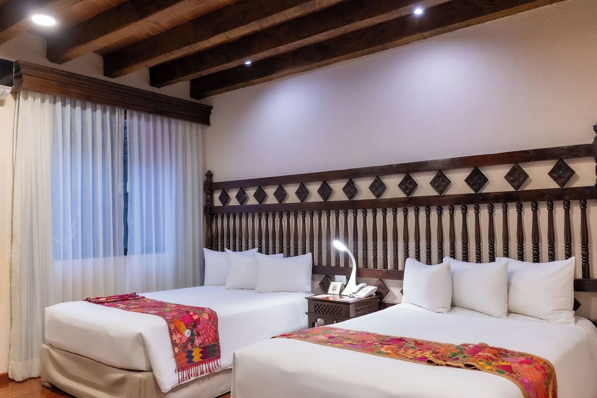 Habitación de hotel con dos camas, vigas de madera en el techo, una araña y un ventanal con cortinas.