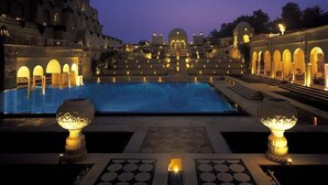 A large, illuminated swimming pool with a central fountain, surrounded by a grand building with arched walkways and a dome structure in the background. - The Oberoi Amarvilas, Agra (Agra)