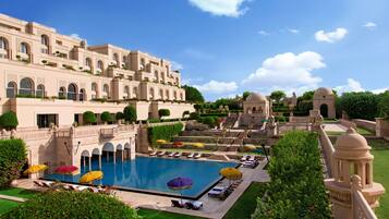 Una gran piscina con tumbonas y sombrillas, rodeada de abundante vegetación y un edificio imponente con múltiples arcos y cúpulas.