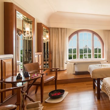 A hotel room with two beds, a sitting area with a chair and table, a bathtub, and large windows with a view of greenery.