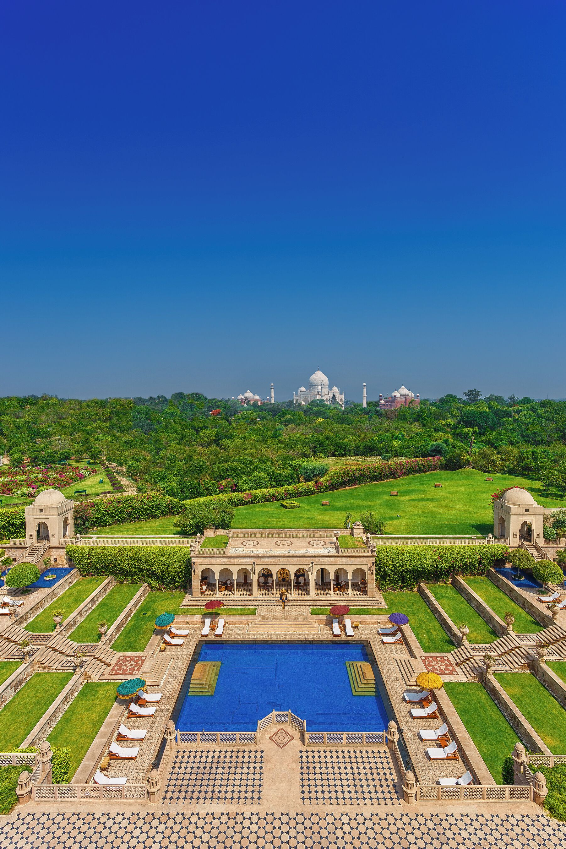A large swimming pool with sun loungers and umbrellas, surrounded by manicured gardens and a view of the Taj Mahal in the distance.