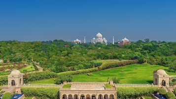 A large swimming pool with sun loungers and umbrellas, surrounded by manicured gardens and a view of the Taj Mahal in the distance.
