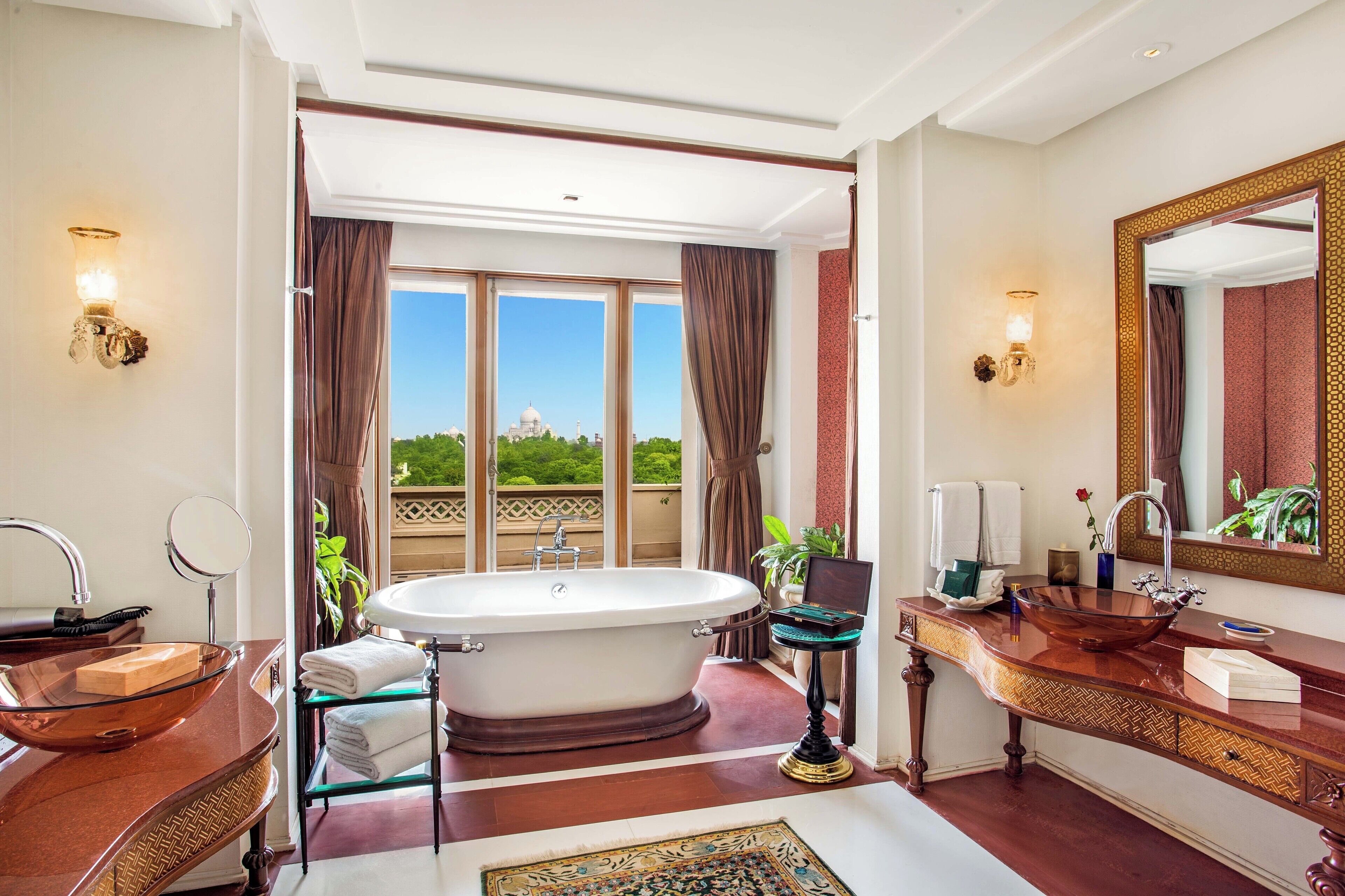 A luxurious bathroom with a freestanding bathtub, a large mirror, and a view of the Taj Mahal through large windows.