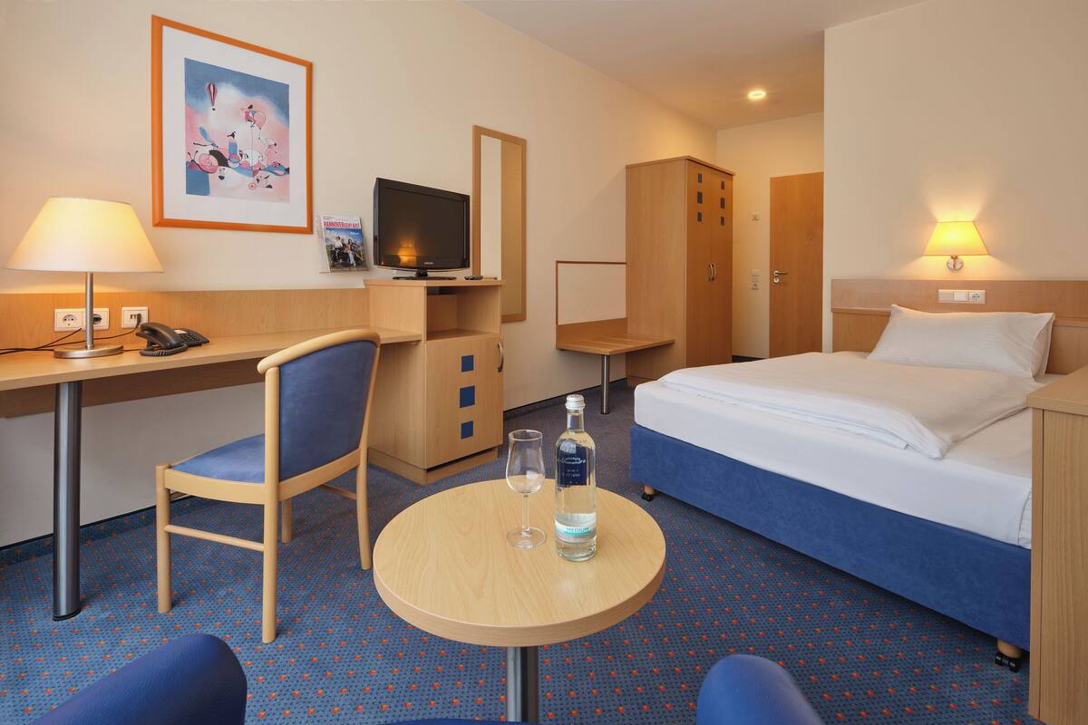 A hotel room with a bed, a desk with a chair, a small table with a glass and bottle, a TV, and a mirror.