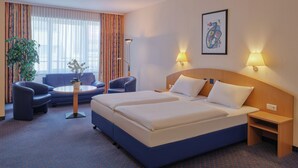 A hotel room with a large bed, two bedside tables, a desk with a chair, a sofa, and a potted plant. - YORS Hotel Hannover (Hannover)