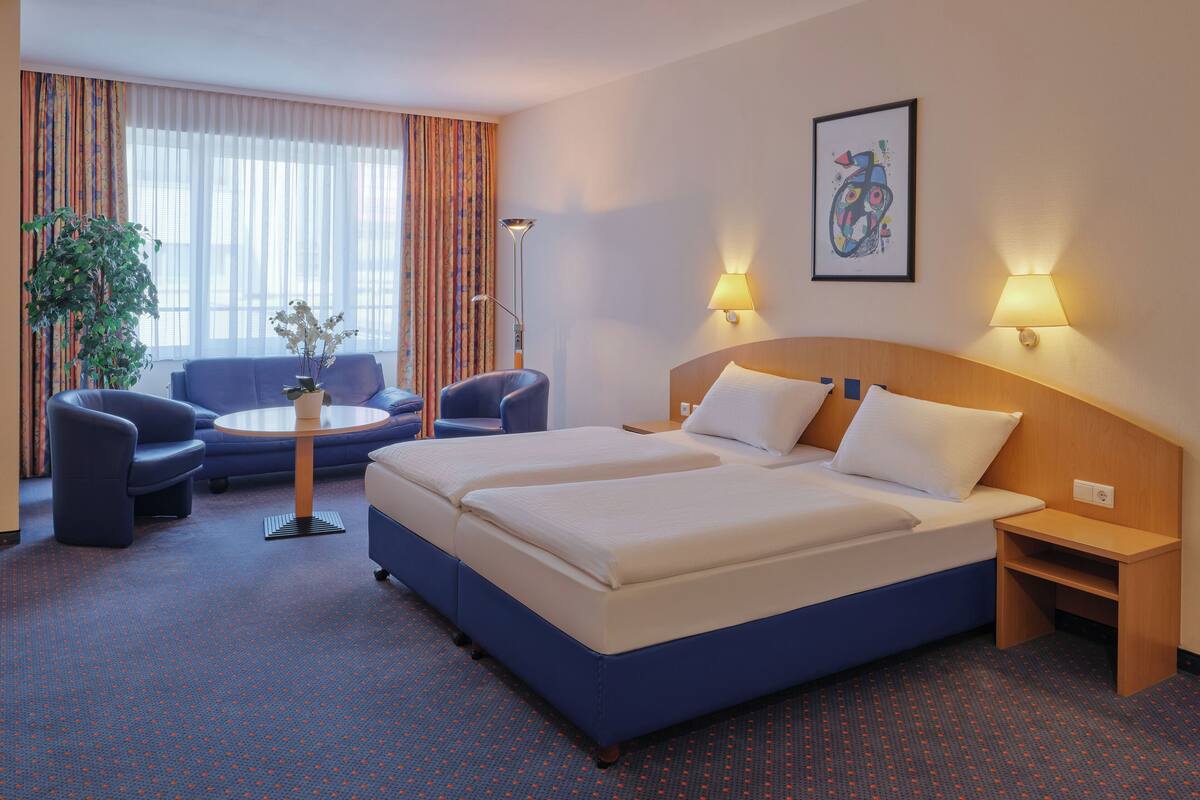 A hotel room with a large bed, two bedside tables, a desk with a chair, a sofa, and a potted plant.