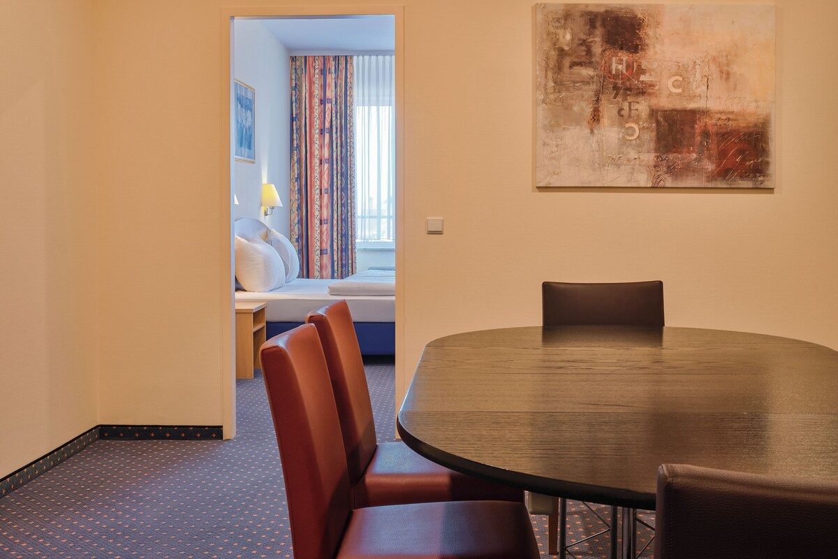 A hotel room with a round dining table, red upholstered chairs, a bed with white linens, a colorful curtain, and a large abstract painting on the wall.