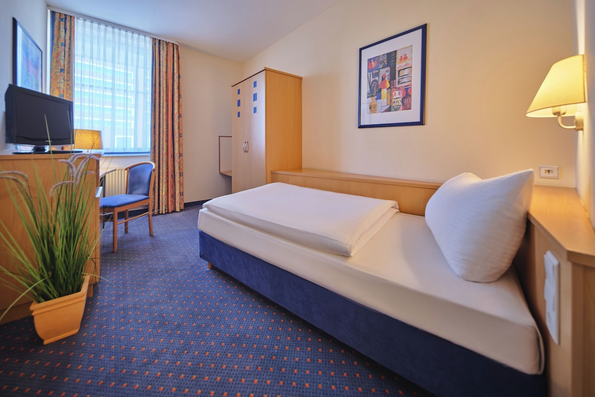 A hotel room with a bed, a desk with a computer, a chair, a wardrobe, and a potted plant.