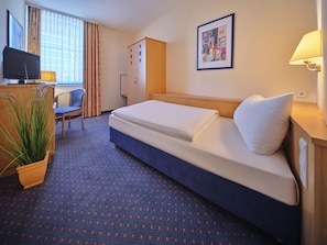 A hotel room with a bed, a desk with a computer, a chair, a wardrobe, and a potted plant. - YORS Hotel Hannover (Hannover)