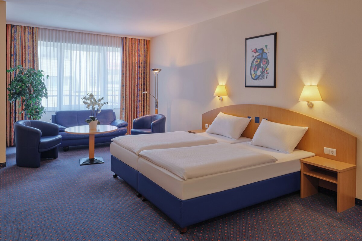 A hotel room with a large bed, two bedside tables, a desk with a chair, a sofa, and a potted plant.