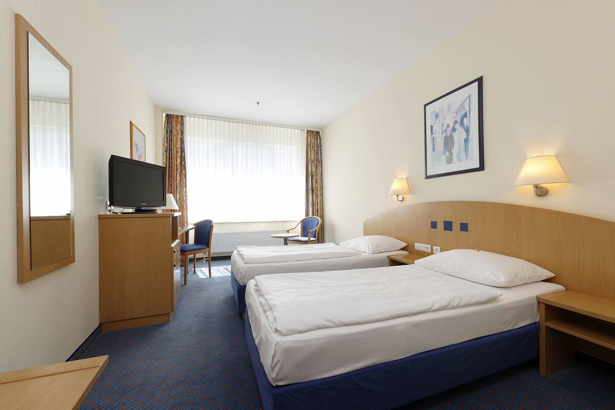 A hotel room with two beds, a desk with a TV, a chair, and a window with curtains.