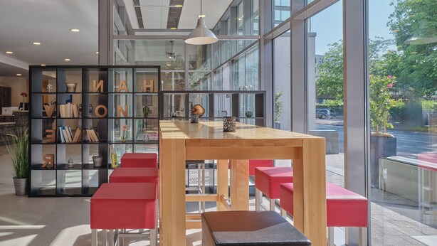 A modern interior with a wooden table and red chairs, large windows, and a bookshelf with letters. - YORS Hotel Hannover (Hannover)