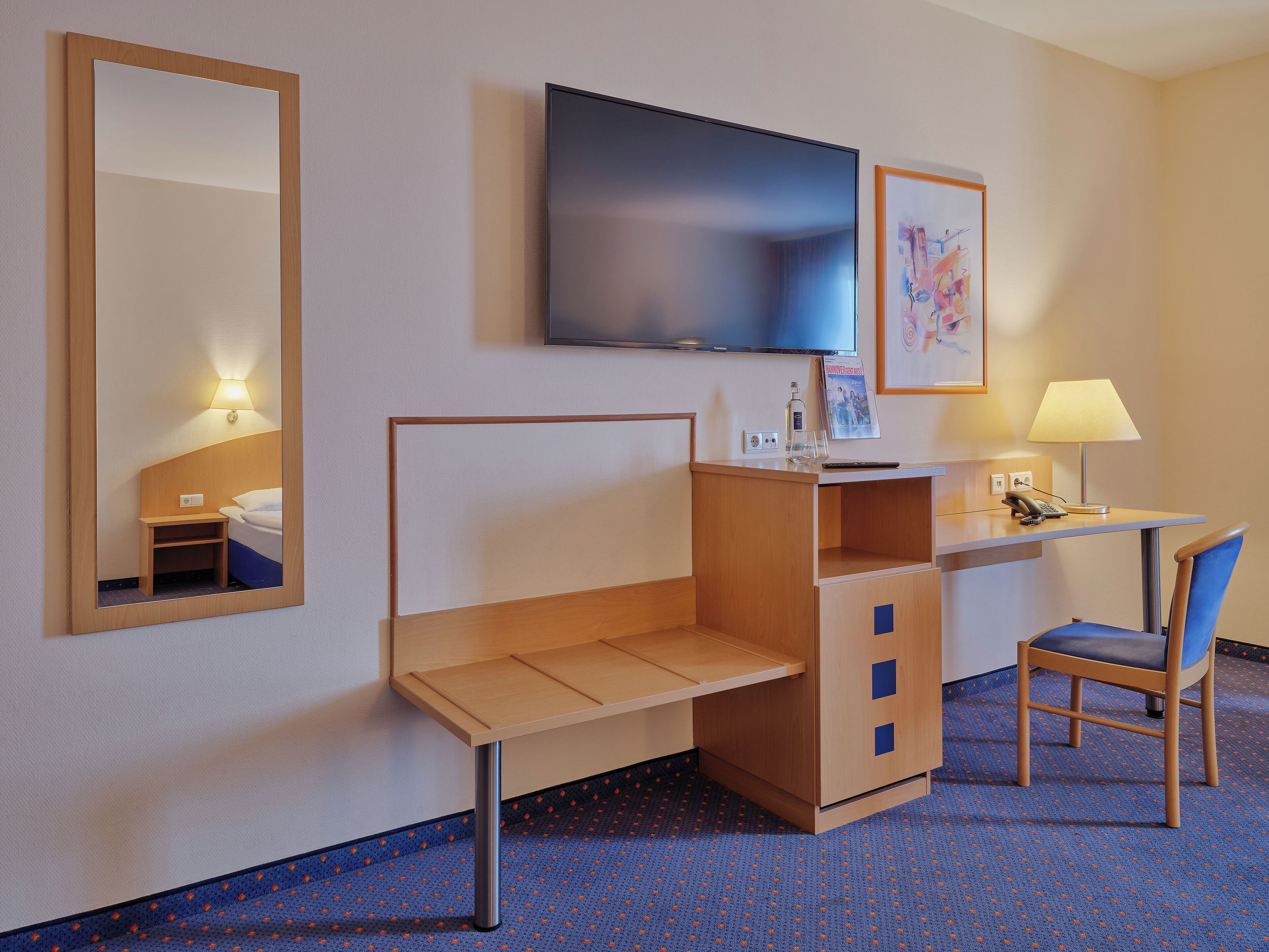 A hotel room with a bed, a desk with a lamp, a chair, a TV, and a mirror.
