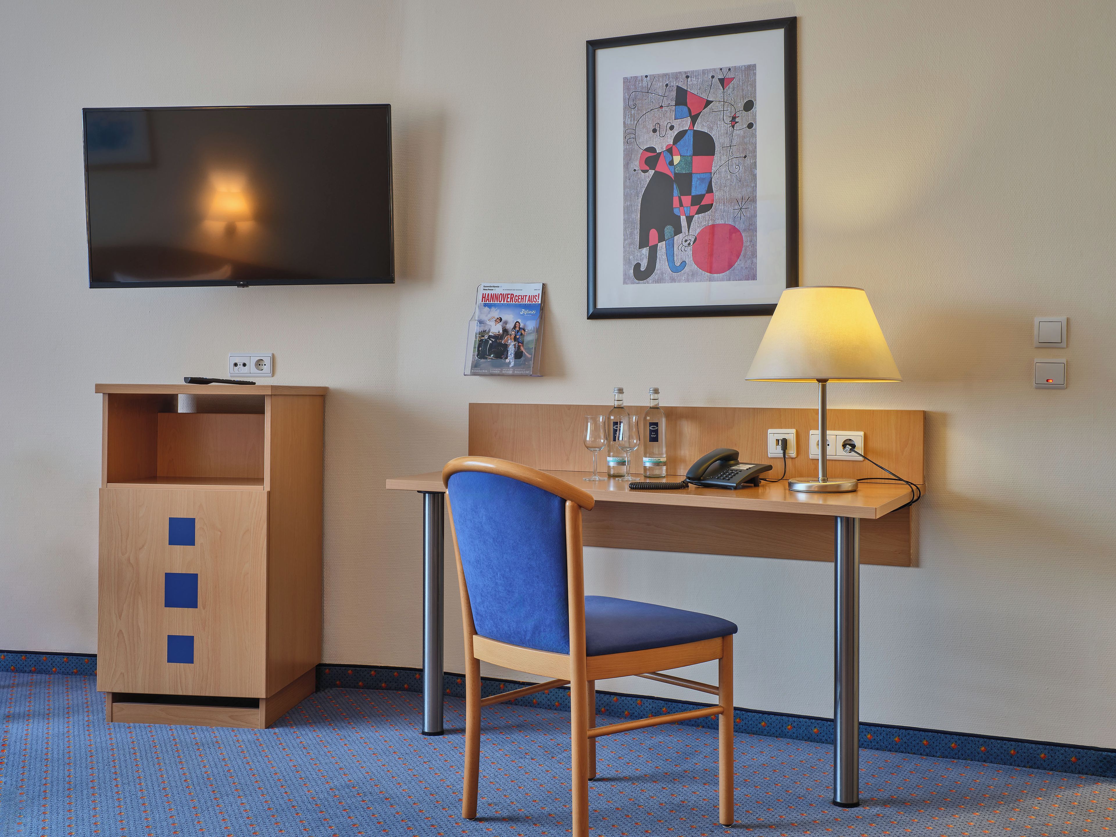 Ein Hotelzimmer mit Schreibtisch, Stuhl, Fernseher, gerahmten Bildern und einer Lampe.