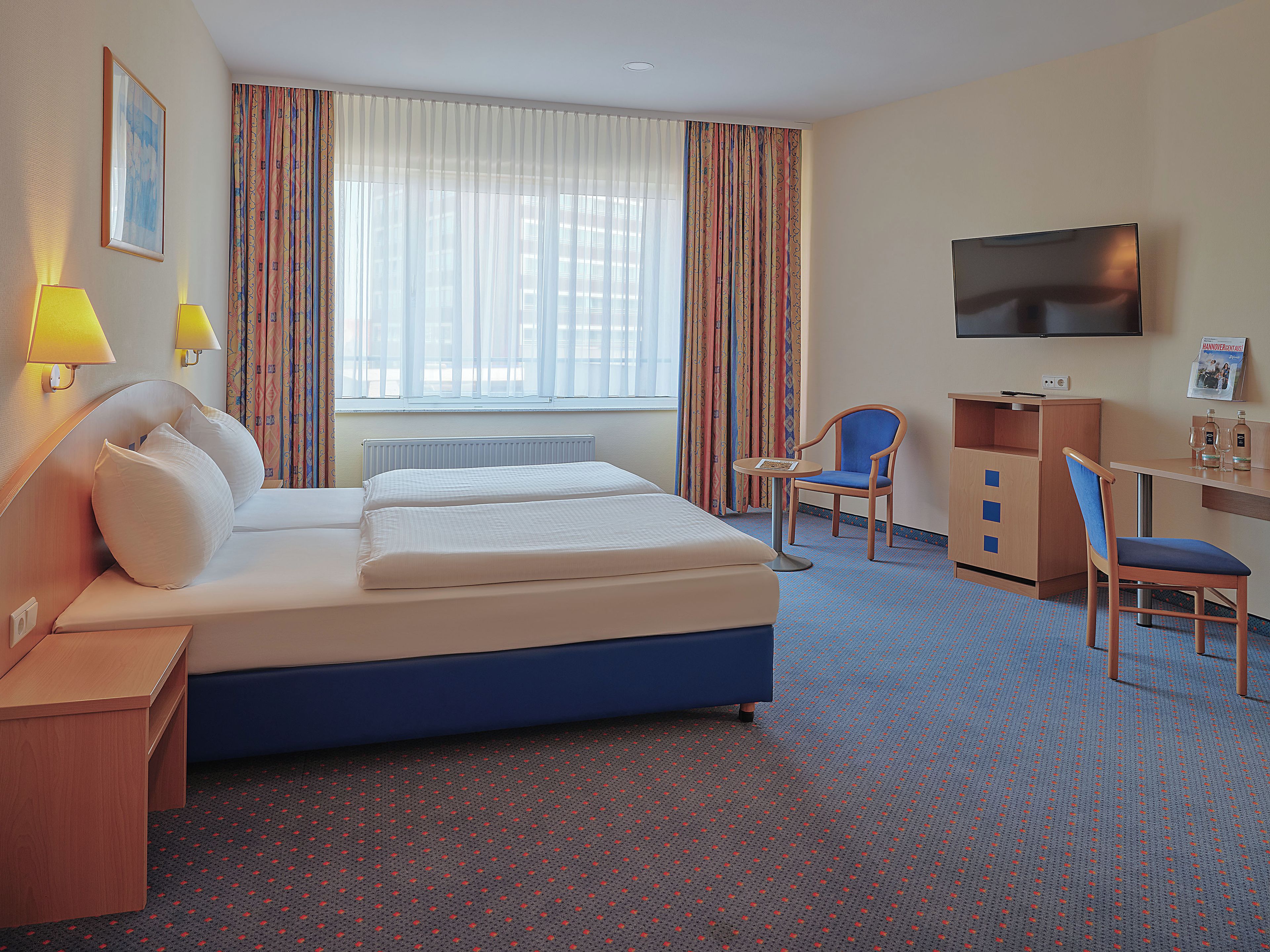 A hotel room with a bed, a desk, a chair, a TV, and a window with curtains.