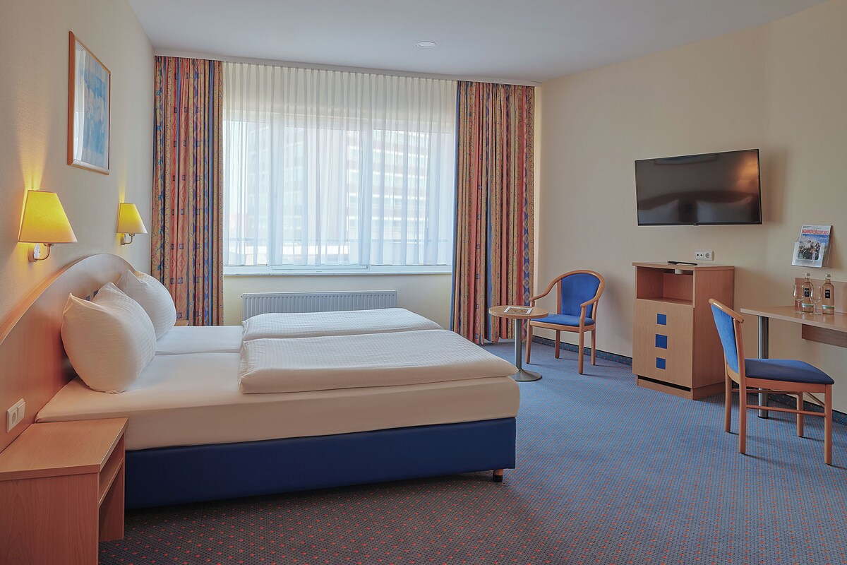 A hotel room with a bed, a desk, a chair, a TV, and a window with curtains.