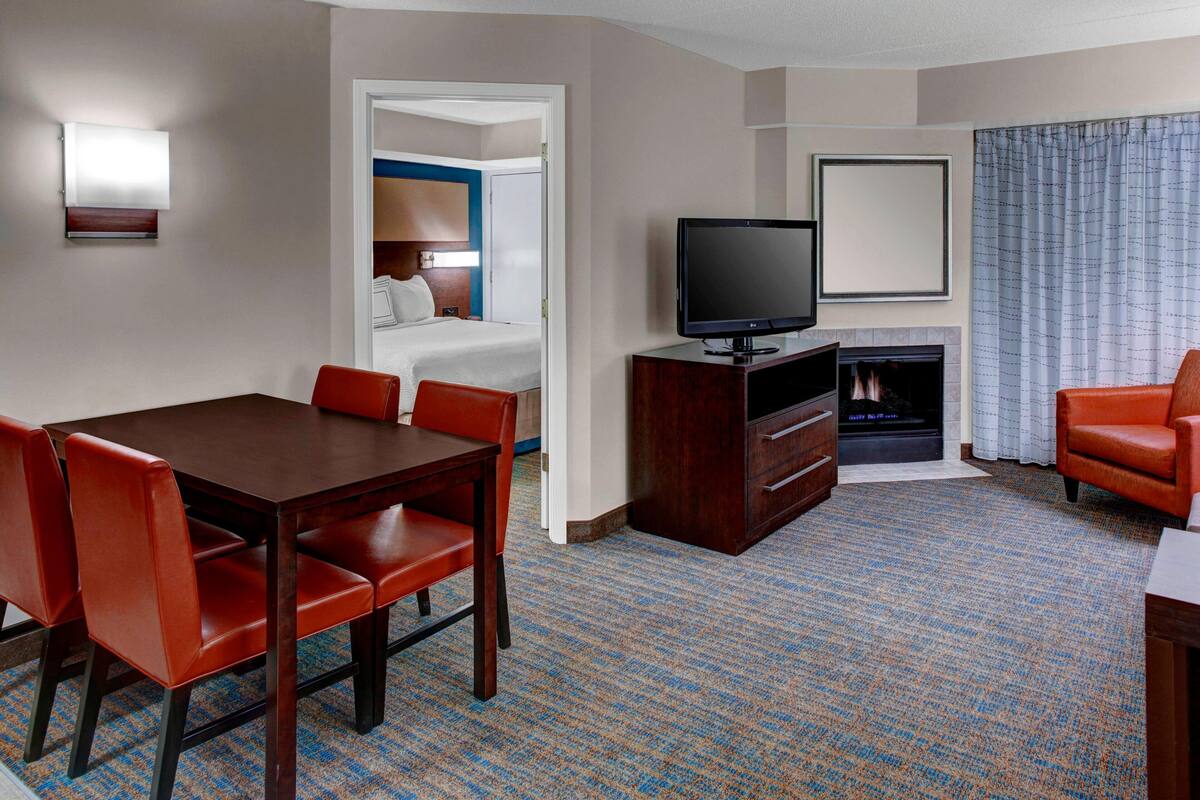 A hotel room with a dining table, red chairs, a flat-screen TV, a fireplace, and a large window with curtains.