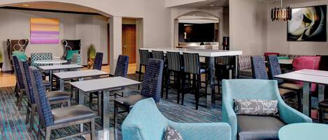 A hotel lobby with a mix of seating options including sofas and chairs, a central dining area with tables and chairs, and a large screen TV mounted on the wall.