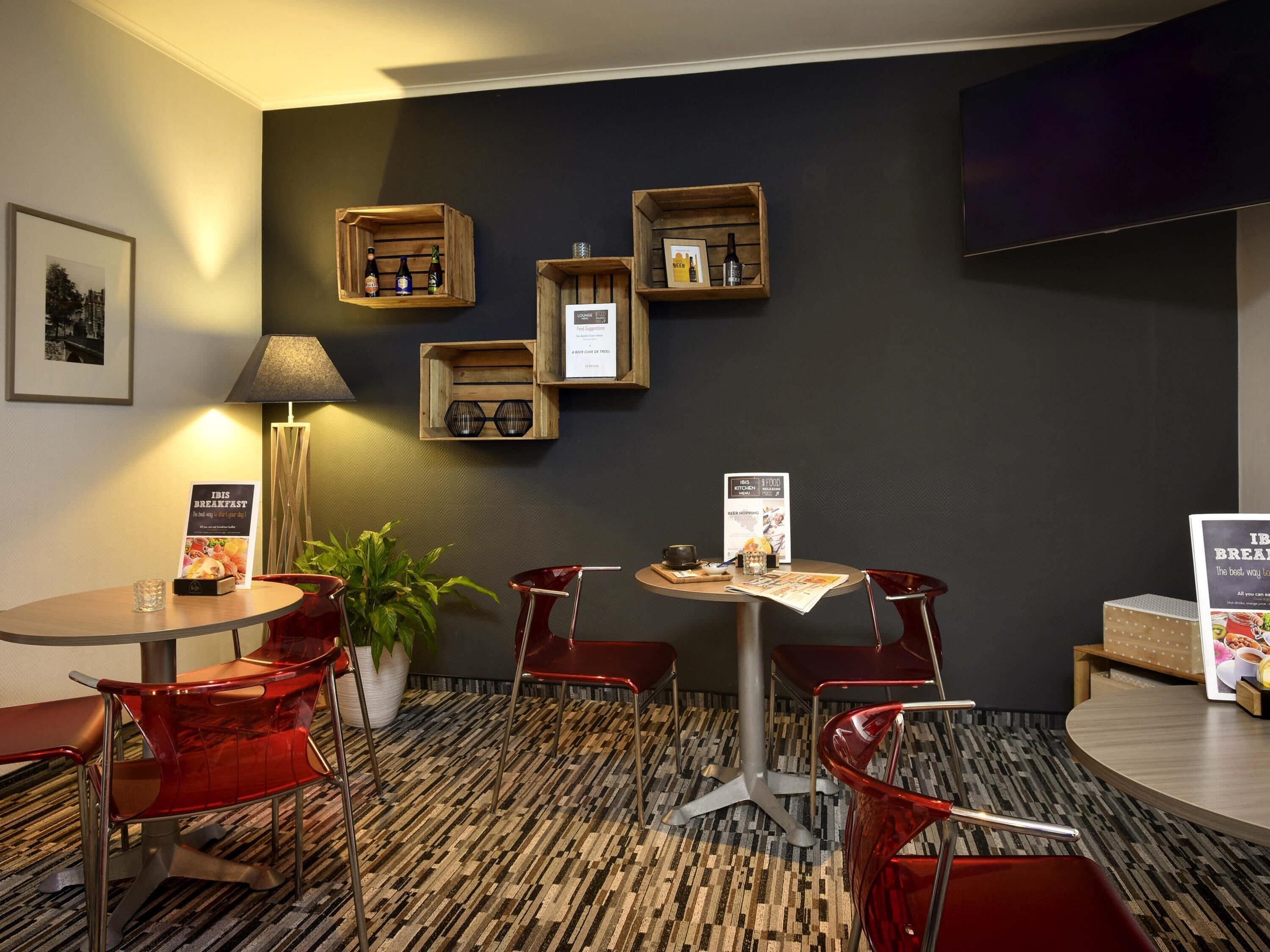 Habitación de hotel con una mesa de comedor redonda, sillas rojas, una planta en maceta y estantes en la pared con varios objetos.