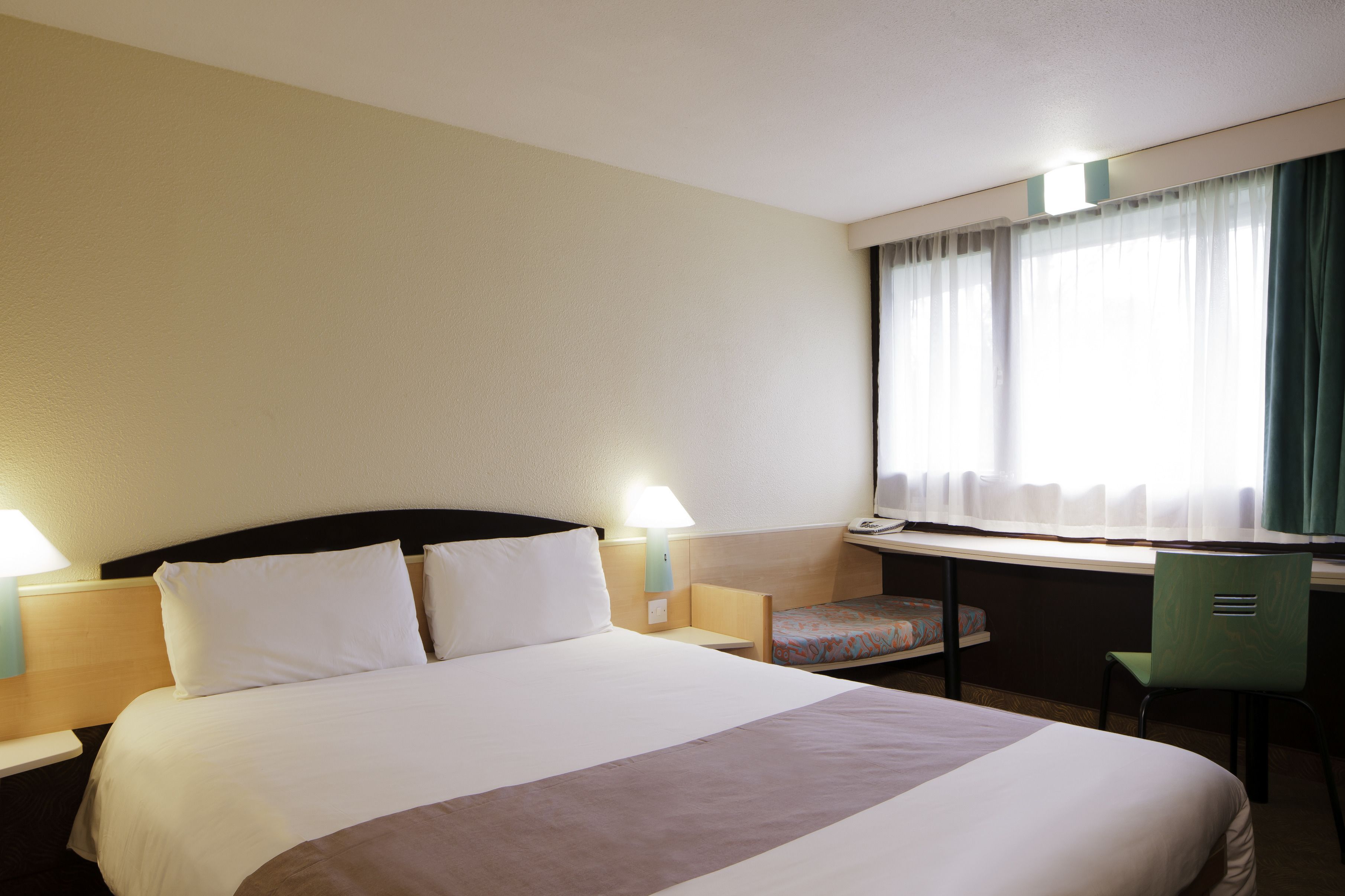 A hotel room with a large bed, a desk with a chair, and a window with curtains.