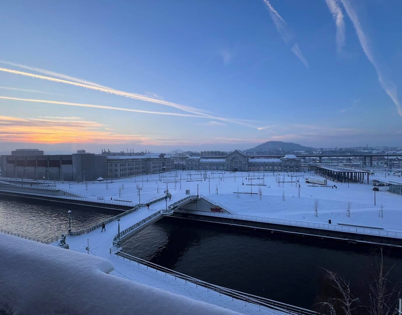 A snow-covered outdoor area with a water body, buildings, and a distant cityscape.