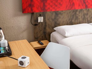 A hotel room with a wooden desk, a white power strip, a remote control, a bed with white bedding, and a red accent wall. - ibis Coventry South (Coventry)