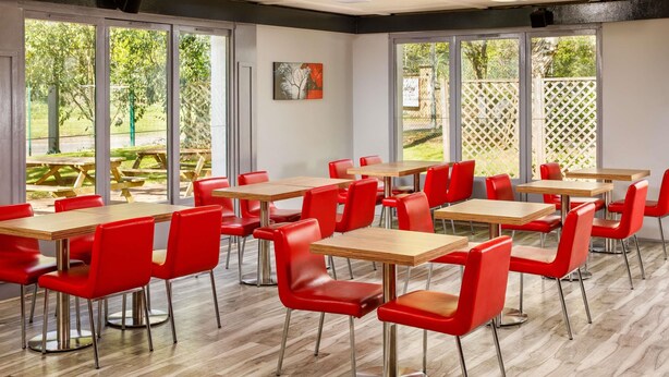 A dining area with multiple tables and red chairs, large windows, and a view of an outdoor patio. - ibis Coventry South (Coventry)