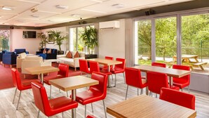 A modern indoor space with multiple red chairs and wooden tables, a sofa, and a TV on the wall. - ibis Coventry South (Coventry)