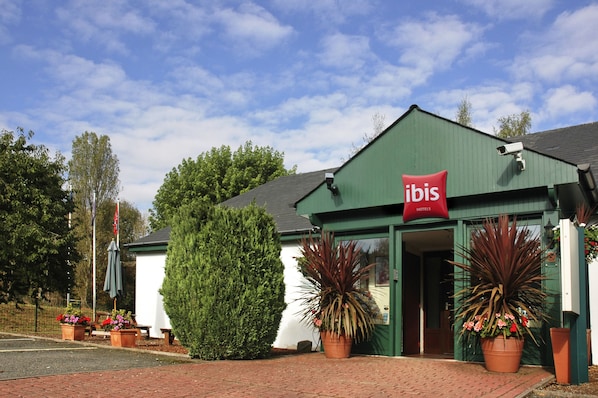 Un bâtiment vert avec une enseigne rouge sur laquelle on peut lire « ibis Hotels ». Des plantes en pot sont disposées devant le bâtiment.
