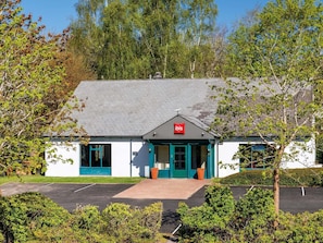 A white building with a red ibis logo, a sloped roof, and a green door. - ibis Coventry South (Coventry)