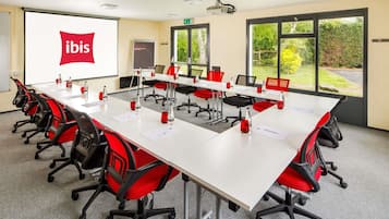 Una sala de conferencias con una larga mesa, sillas rojas y negras, una pantalla de proyector y vistas al exterior con vegetación.