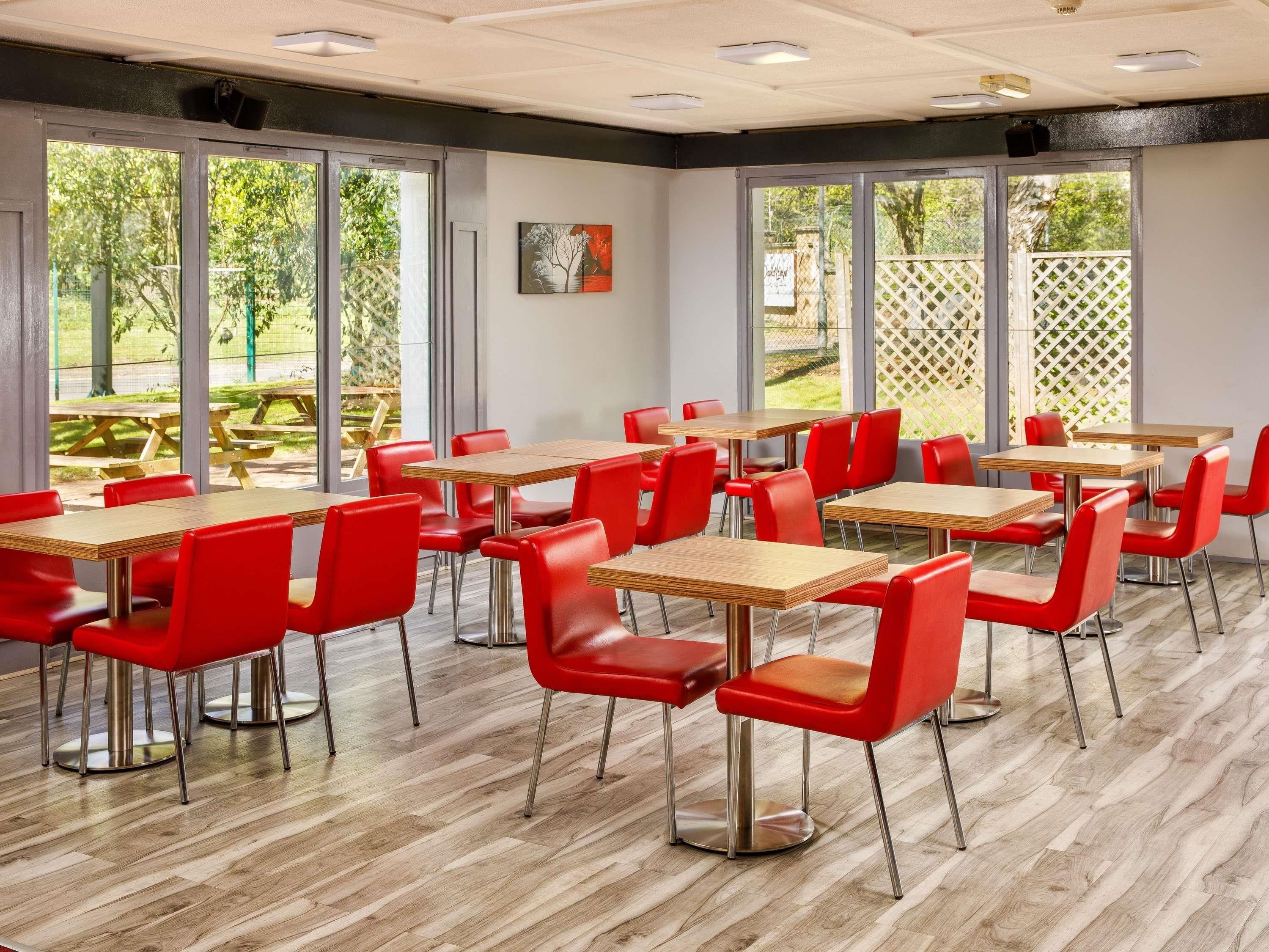 Comedor con varias mesas y sillas rojas, ventanas amplias y vistas a un patio exterior.