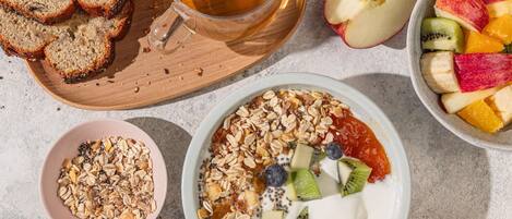 Frühstücksszene mit Brot, einer Tasse Tee, einer Obstschale, einer Schale Granola und einem Teller Joghurt mit Früchten.