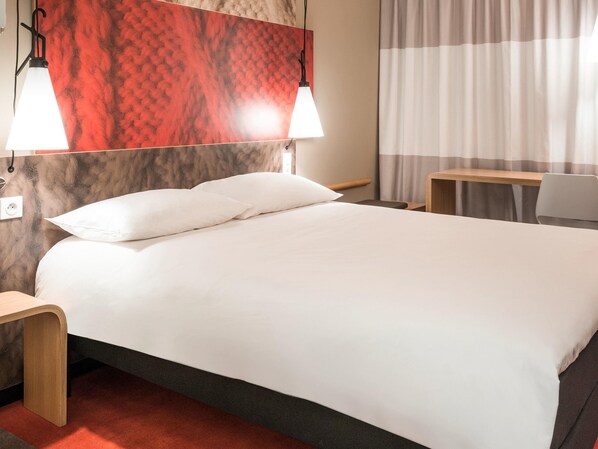 A hotel room with a bed, two bedside lamps, a wooden nightstand, a desk, a chair, and a window with curtains.