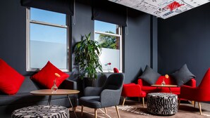 A modern lounge area with a grey sofa, red armchairs, a round wooden table, and patterned ottomans. - ibis Coventry Centre (Coventry)