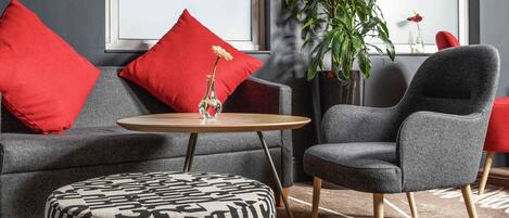 Una sala de estar moderna con un sofá gris, cojines rojos, una mesa redonda de madera, un sillón gris y un reposapiés estampado.