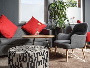 A modern living room with a grey sofa, red cushions, a small round table, a grey armchair, and a patterned ottoman. - ibis Coventry Centre (Coventry)