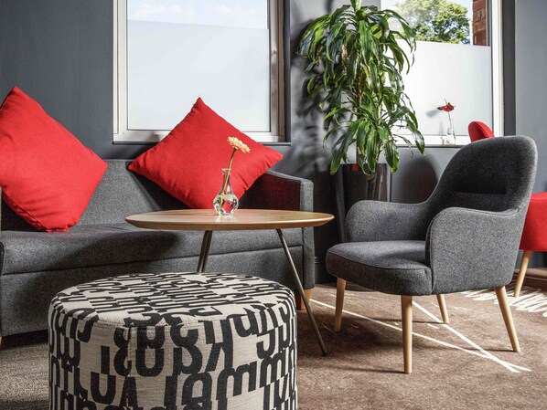 A modern living room with a grey sofa, red cushions, a small round table, a grey armchair, and a patterned ottoman. - ibis Coventry Centre (Coventry)