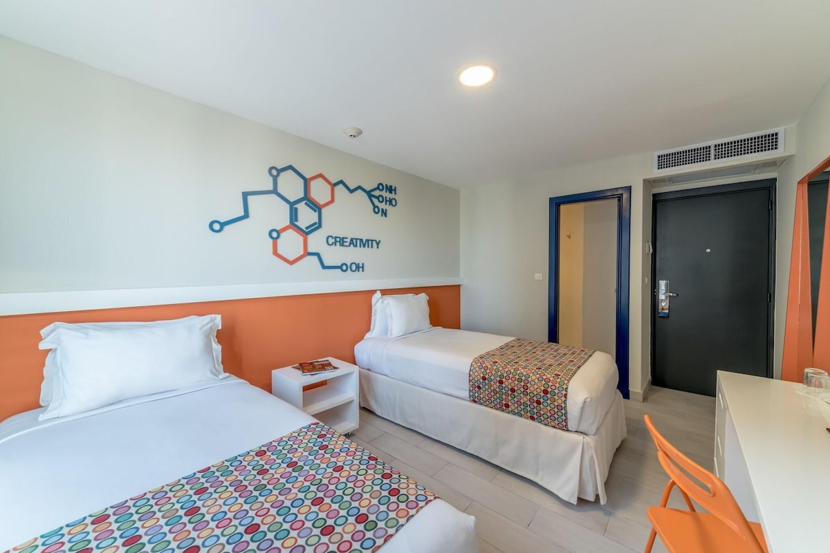 A hotel room with two beds, a desk, and a chair. There is a wall decoration with a molecular structure and the word "CREATIVITY".
