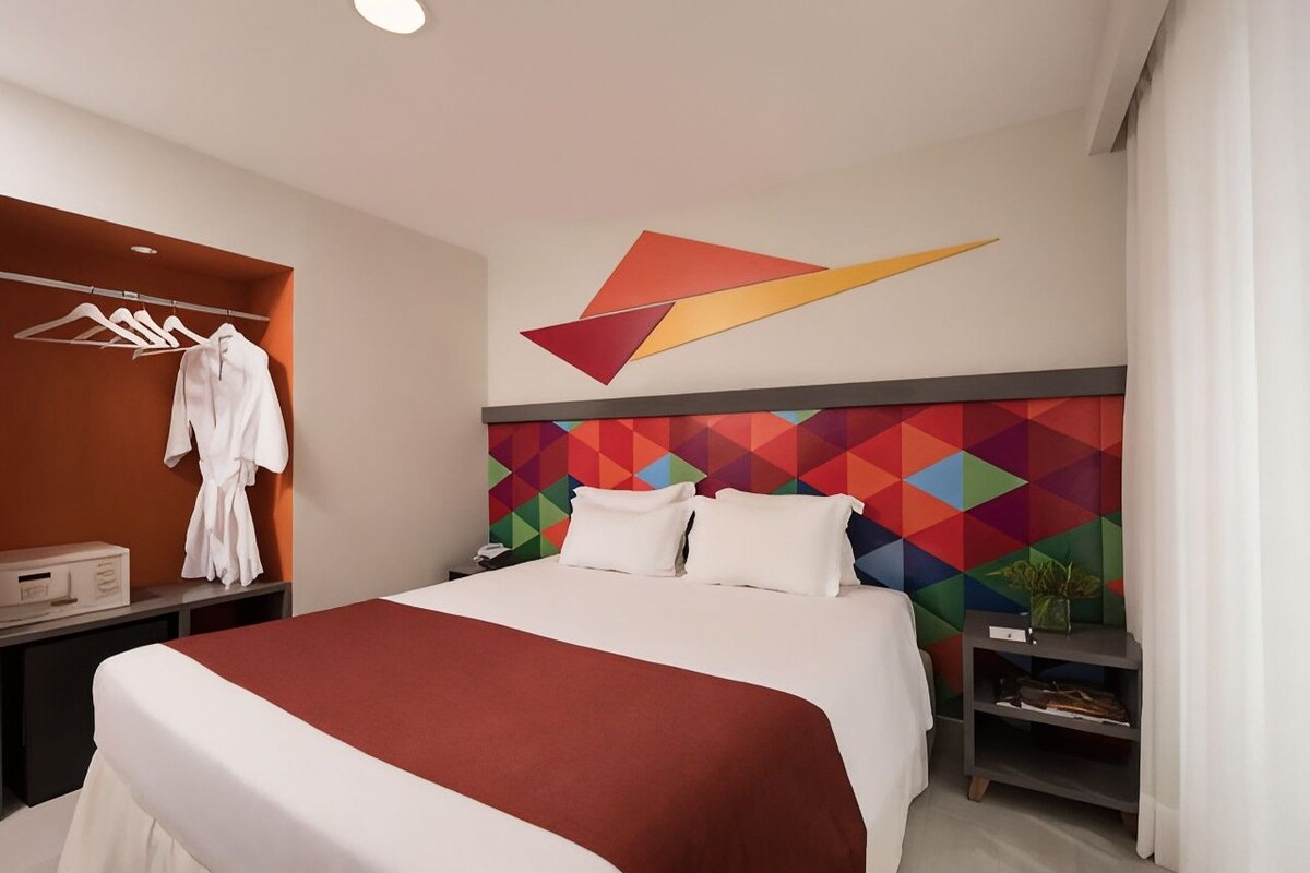 Habitación de hotel con cama, perchero, mesita de noche y un cabecero con estampado geométrico.