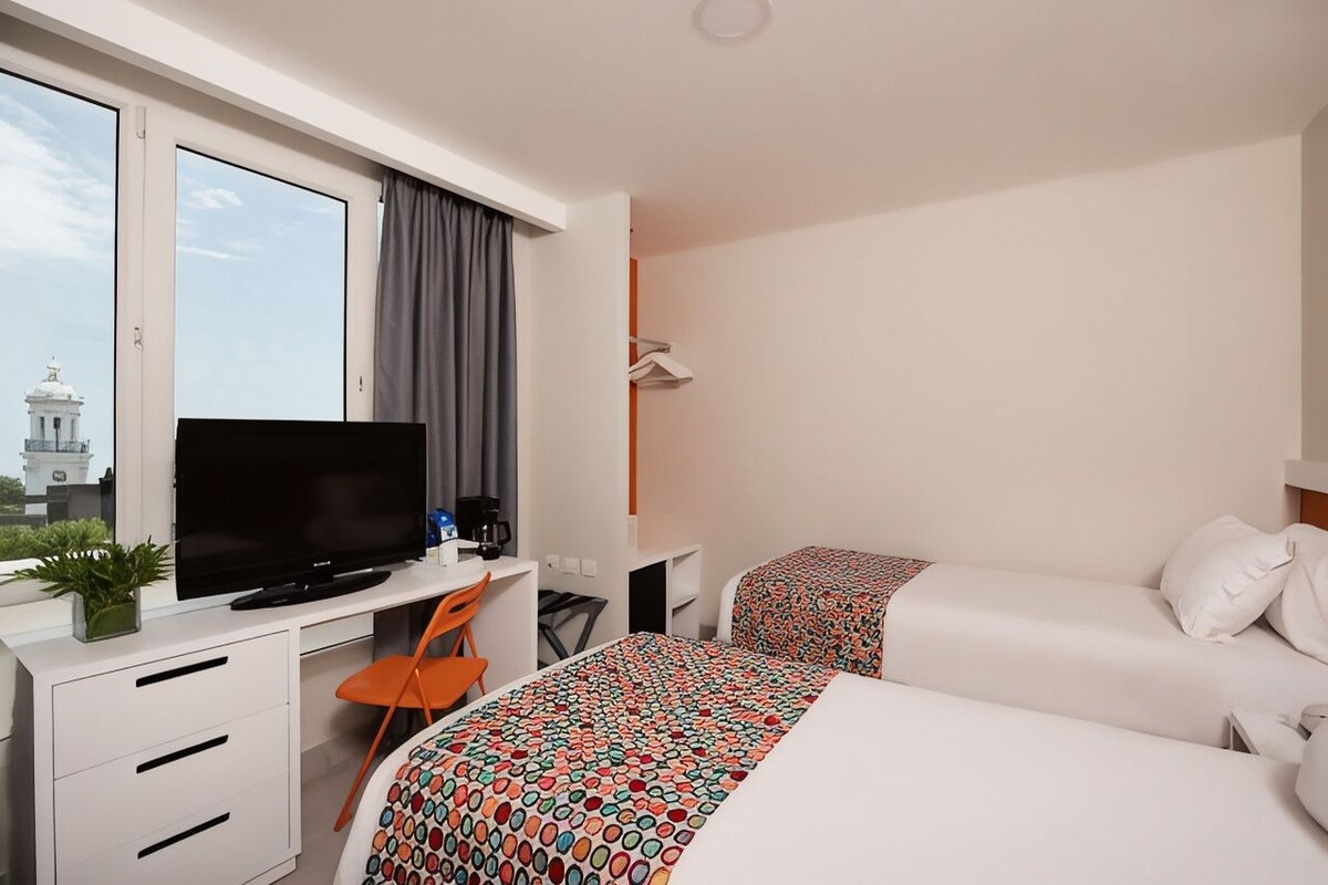 Habitación de hotel con cama, escritorio con televisor, una silla y ventana con cortinas.